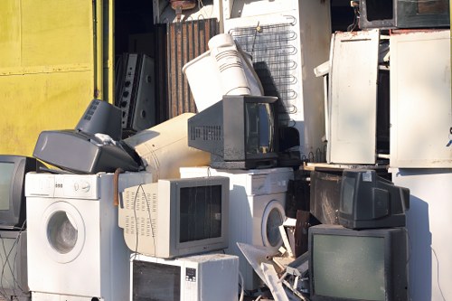 A collection of discarded household appliances and electronic devices piled together outdoors, including washing machines, microwave ovens, and small refrigerators with varied finishes such as white, black, and metallic silver. The appliances are stacked and leaning against each other on a paved surface, situated in front of a warehouse or storage shed with corrugated metal walls in the background. Some items show signs of dirt and wear, and the scene is illuminated by natural daylight. The setting suggests a waste collection or removal area, consistent with services offered by Commercial Waste Removal Mortlake in the local area, possibly near the TW9 postcode. The arrangement of waste appliances highlights the importance of professional rubbish removal to responsibly manage bulky household trash.