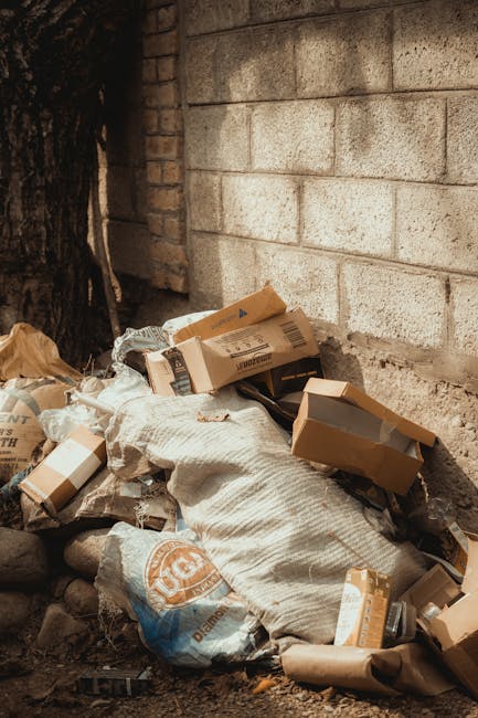 Close-up view of a person wearing an orange protective suit and white gloves, holding a large blue plastic rubbish bag tightly by the top. The bag is filled with waste and appears to be secured for di