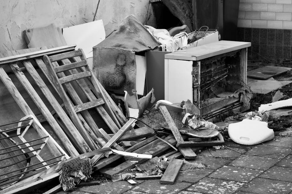 A collection of discarded household items and debris positioned against an outdoor wall, including a wooden landscaped garden stool with horizontal slats, positioned on the left side of the image. Nex