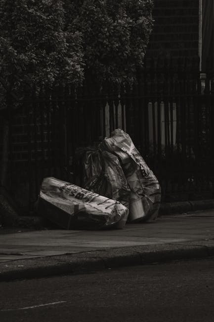 Two large black plastic rubbish bags, filled and tied at the top, are positioned on a sidewalk next to a curb in front of a black metal fence with vertical bars. One bag appears slightly larger and is