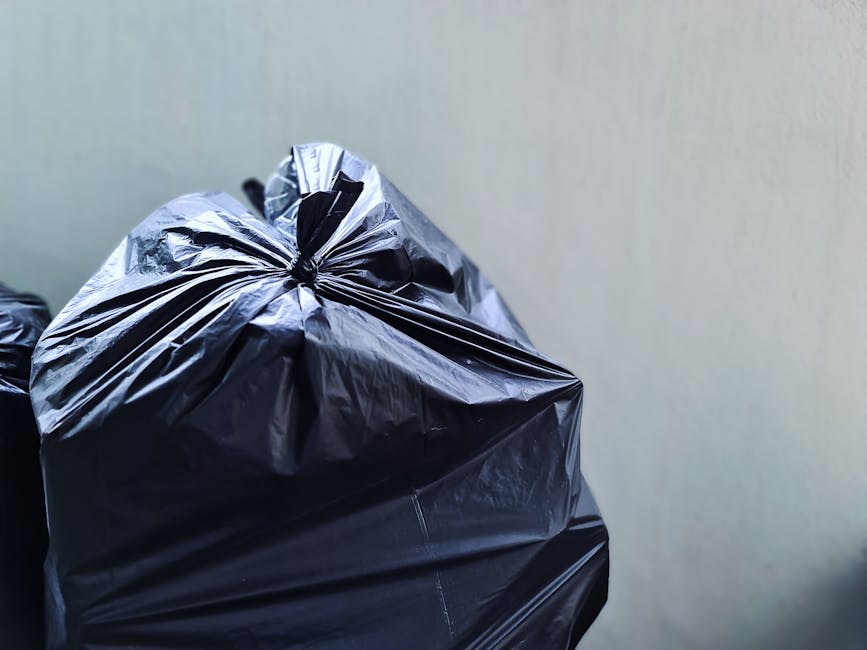 Two large black plastic rubbish bags, filled and tied at the top, are positioned on a sidewalk next to a curb in front of a black metal fence with vertical bars. One bag appears slightly larger and is