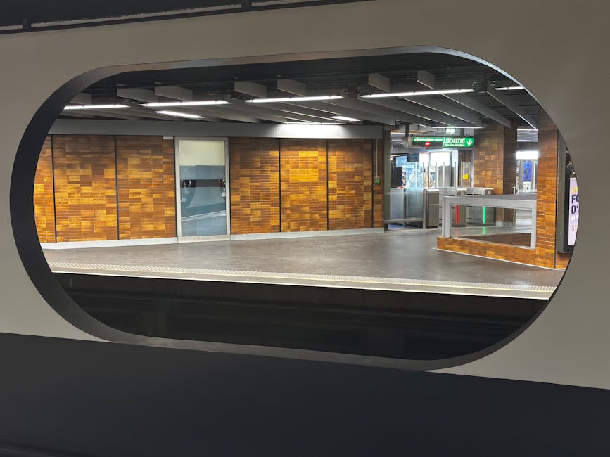 View of a modern underground station platform framed by an oval window, featuring a wall clad in warm-toned wooden panels arranged in a rectangular pattern. The platform surface appears smooth and cle